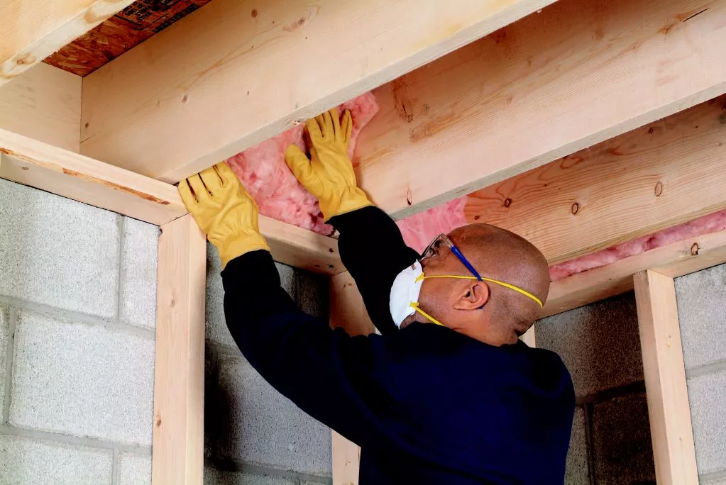 Technician installing pink fiberglass insulation in a crawl space ceiling while wearing a white face mask and yellow gloves.